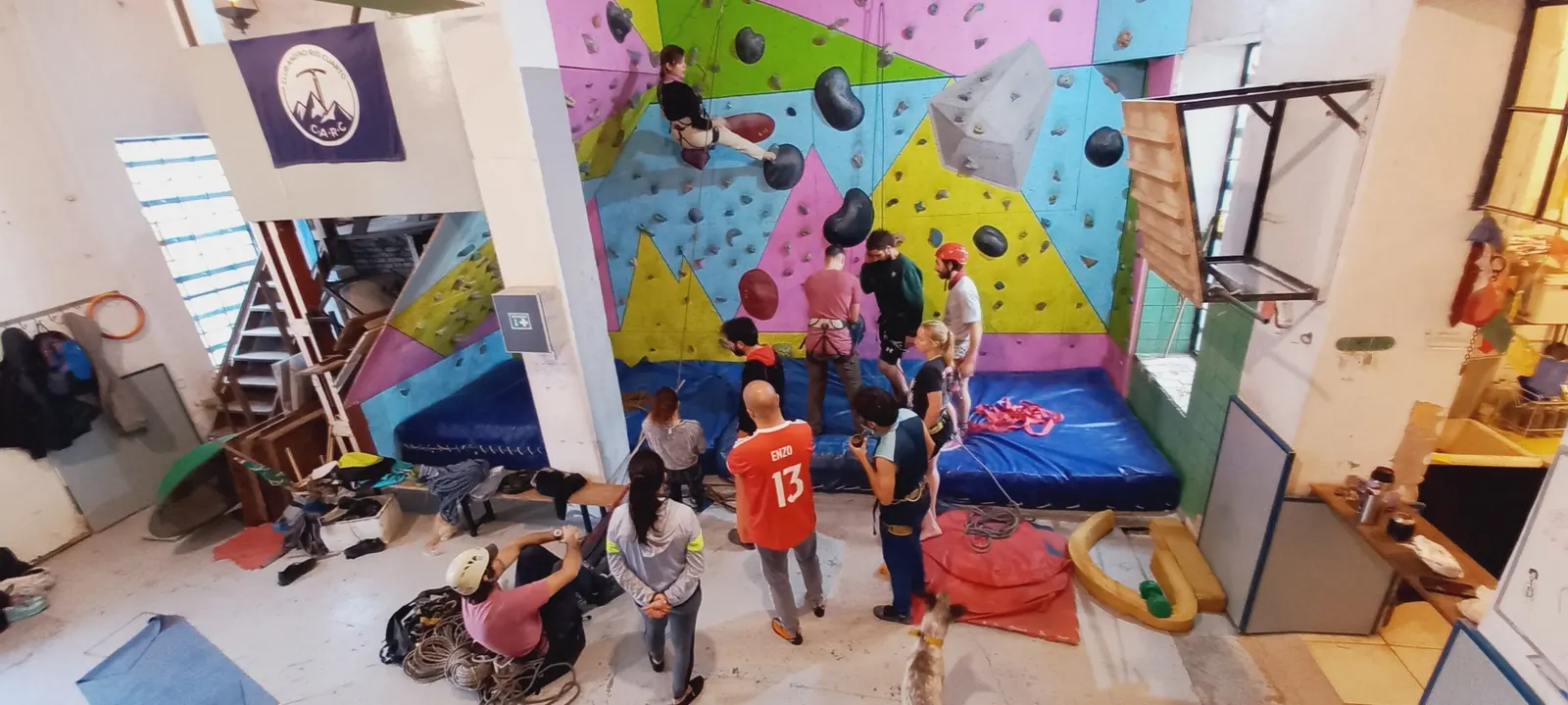 Clase en el muro de escalada del Club Andino Río Cuarto: un grupo de alumnos y profesores al pie de un muro de colores, con la bandera del club colgada a la izquierda